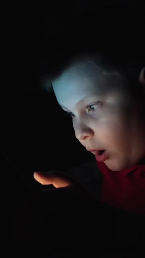 A boy is playing on a tablet in complete darkness, lying on a bed. Video stock 267647043