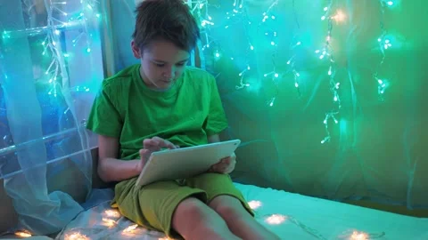 A boy is playing a tablet sitting on a bed decorated with garlands. multicolored Stock Footage 170783991