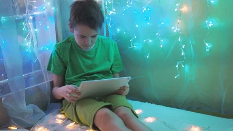 A boy is playing a tablet sitting on a bed decorated with garlands. multicolored Stock Footage 172070129
