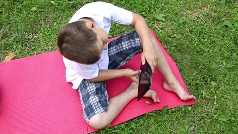 A boy is playing on a tablet while sitting on the grass in the park Stock Footage 92153478
