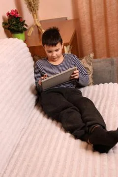 A boy plays games on a tablet while sitting on the couch. Stock Photos