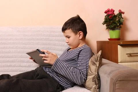 A boy plays games on a tablet while sitting on the couch. Stock Photos