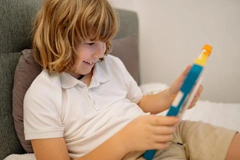 Boy plays on tablet while sitting on bed in his bedroom during the day Stock-Fotos