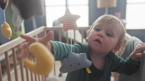Boy reaching out hand to grab hanging plush mobile above wooden crib in bui.. Stock Footage 339236715