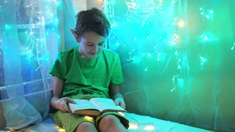 A boy is reading a book lying on the sofa. into the light of the garlands Stock Footage 171629391