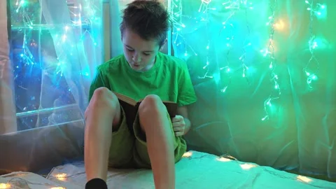 The boy is reading a book sitting on the bed in the light from the garlands. the Stock Footage 170072553