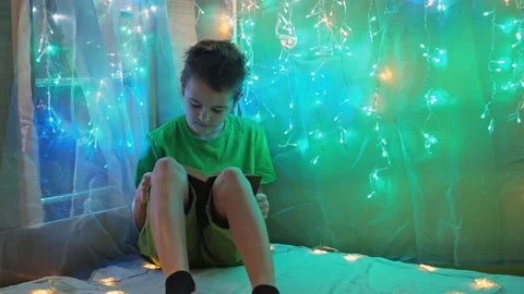 The boy is reading a book sitting on the bed in the light from the garlands. the Stock Footage 170247636