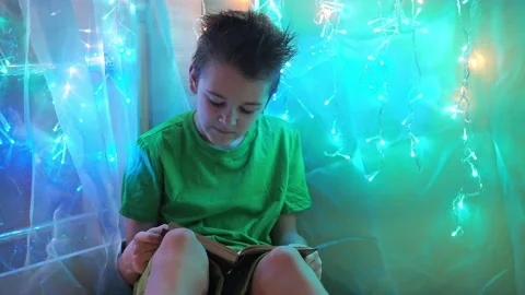 The boy is reading a book sitting on the bed in the light from the garlands. the Stock Footage 170716803