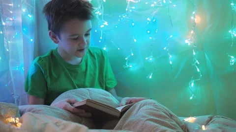 A boy is reading a book sitting on a bed decorated with garlands. multicolored Stock Footage 170716870
