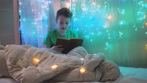 A boy is reading a book sitting on a bed decorated with garlands. multicolored Stock Footage 171012520