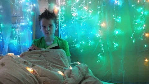 A boy is reading a book sitting on a bed decorated with garlands. multicolored Stock Footage 171129795