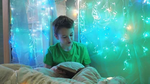 A boy is reading a book sitting on a bed decorated with garlands. multicolored Stock Footage 171470274