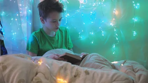 A boy is reading a book sitting on a bed decorated with garlands. multicolored Stock Footage 171629280