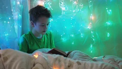 A boy is reading a book sitting on a bed decorated with garlands. multicolored Stock Footage 172154379