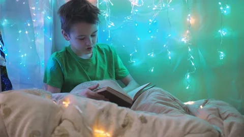 A boy is reading a book sitting on a bed decorated with garlands. multicolored Stock Footage 172257361