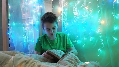 A boy is reading a book sitting on a bed decorated with garlands. multicolored Stock Footage 180115954