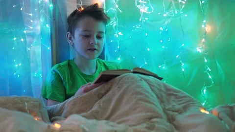 A boy is reading a book sitting on a bed decorated with garlands. multicolored Stock Footage 181524145