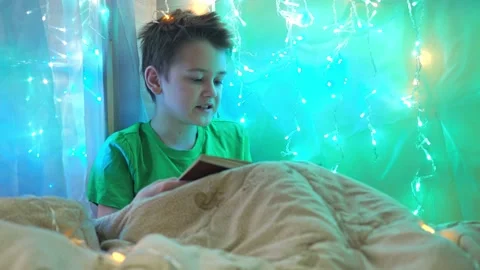 A boy is reading a book sitting on a bed decorated with garlands. multicolored Stock Footage 186126433