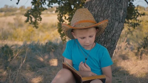 A boy is reading a book sitting under a tree. A child in a straw hat reads. Stock Footage 120707883
