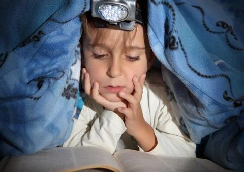 Boy reading a book under the covers with a flashlight Stock Photos