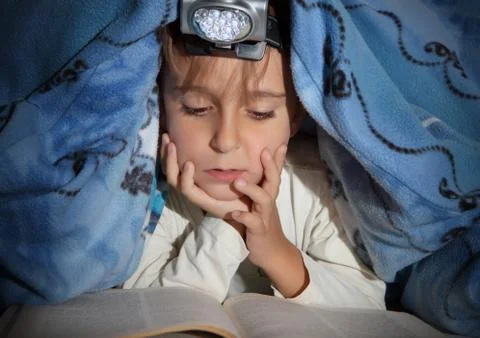 Boy reading a book under the covers with a flashlight Stock Photos