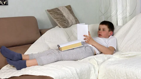 Boy reading paper book aloud, lying on sofa at home, studying at home. Stock Footage 154309746