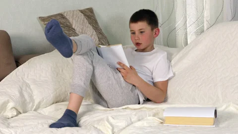 Boy reading paper book aloud, sitting on sofa, studying at home Stock Footage 154309778
