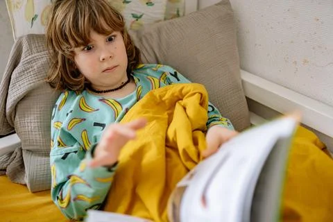 Boy reads a book while sitting in bed wearing pajamas in a cozy bedroom Stock-Fotos