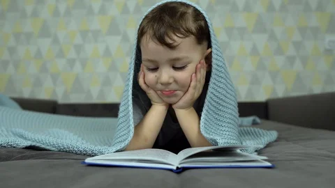 A boy reads an interesting book lying in bed under a blanket Stock Footage 128974706