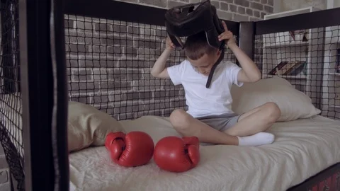 Boy with red Boxing Gloves putting on black Head guard in his bed, ready for Stock Footage 111554299