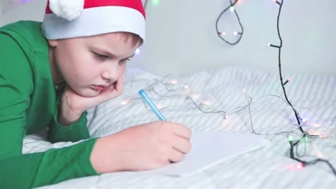 A boy in a red cap of Santa Claus lying on the bed writes a Christmas letter Vídeos de archivo 165384019