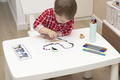 Boy in red pajamas draws with paints at a white table in a home interior Stock Photos