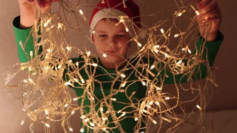 A boy in a red Santa Claus hat is playing with a luminous yellow garland. Stock Footage 165327938
