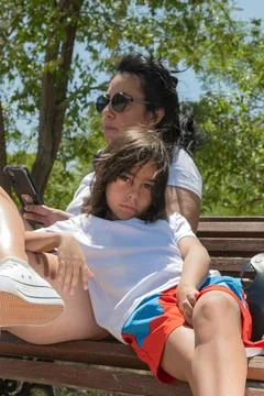 Boy relaxing on bench with mother using smartphone in park Foto stock