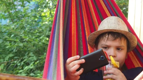 Boy resting in a hammock, drinking a cocktail with lemon, talking on the phone 動画素材 167354920