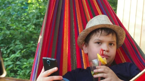 Boy resting in a hammock, drinking a cocktail with lemon, talking on the phone 動画素材 167354927