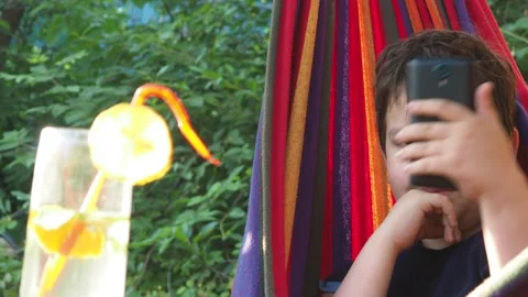 Boy resting in a hammock, drinking a cocktail with lemon, talking on the phone 動画素材 167354960