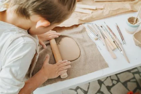 Boy rolling clay piece on a table filled with pottery tools. Stock Photos