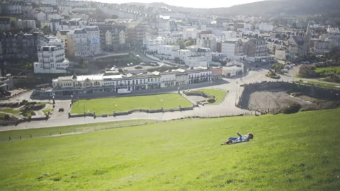 Boy rolls down steep grassy hill towards town Stock Footage 136908245