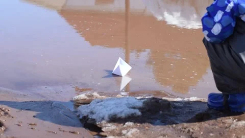 Boy in rubber boots and waterproof pants launches a paper boat into a puddle. Stock Footage 152085340