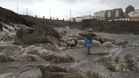 Boy runs around on rocks on dark beach in England Stock Footage 136908473