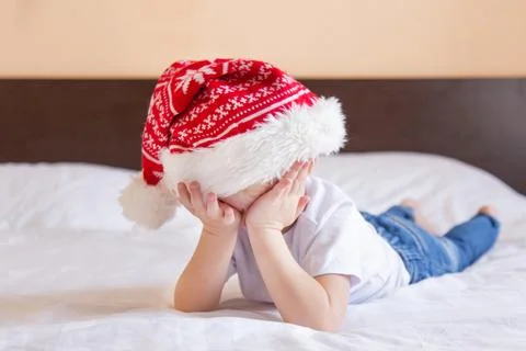 Boy in a santa hat lying on the bed covers his face with his hands. child tired Stock Photos