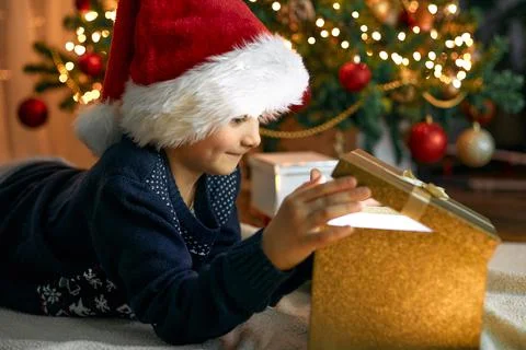 A boy in a santa hat opens a gift box next to a garlanded Christmas tree. Gifts Stock Photos