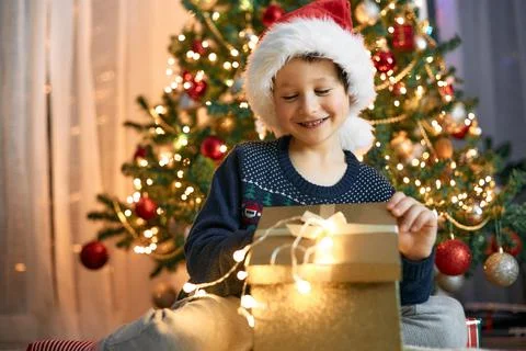 A boy in a santa hat opens a gift box next to a garlanded Christmas tree. Gifts Stock Photos