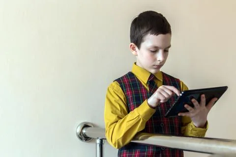 A boy of school age is working on a tablet at the background of light wall. Stock Photos