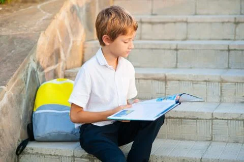 The boy is a schoolboy sitting on the steps of the school and reading a book Stock Photos
