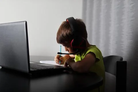 Boy in shadow with headphone writing in notepad next to laptop computer Stock-Fotos
