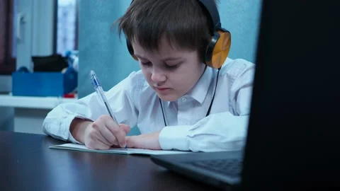 A boy in a shirt writes in a notebook sitting at a laptop Stock Footage 144263365