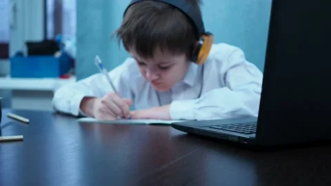 A boy in a shirt writes in a notebook sitting at a laptop. the boy is out of Stock Footage 144684103