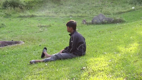 A boy sits alone on the green grass next to the remains of burnt garbage Video stock 338166635